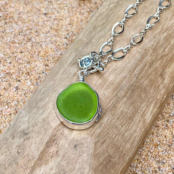 A sea glass pendant necklace with a green sea glass cabochon in a silver bezel setting on a silver chain, displayed on a piece of weathered wood over sand.