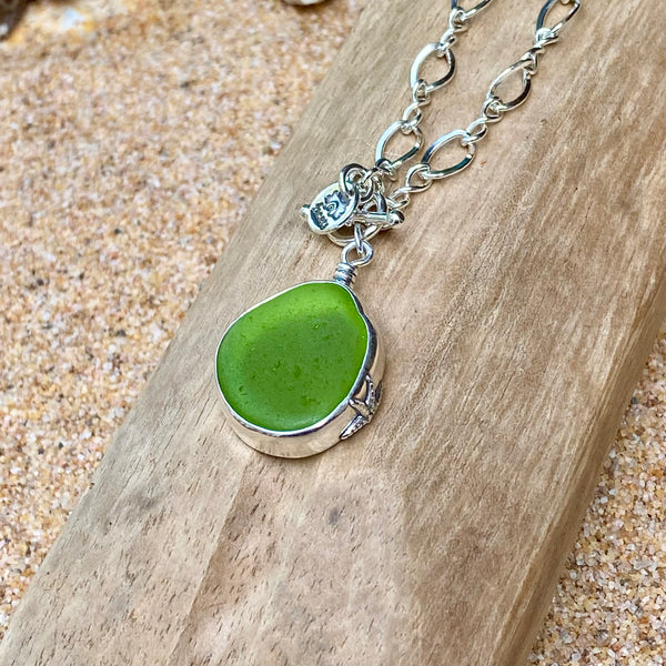 A green sea glass pendant necklace in a silver bezel setting on a silver chain, displayed on a piece of wood over a sandy background.