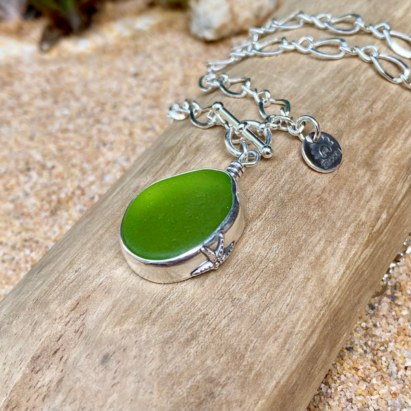 A sterling silver necklace with a polished green sea glass pendant  displayed on a piece of weathered wood over sand.