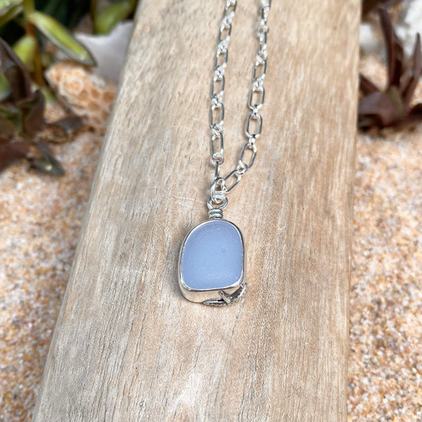 A silver chain necklace with a milky cornflower blue sea glass pendant, displayed on a piece of weathered wood over sand.
