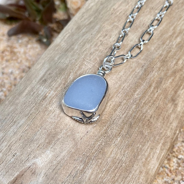 A silver chain necklace with a milky cornflower blue sea glass pendant, displayed on a piece of weathered wood over sand.