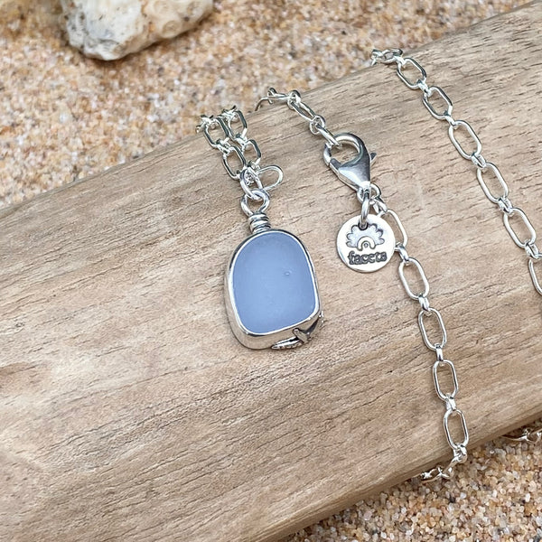 A silver chain necklace with a milky cornflower blue sea glass pendant, displayed on a piece of weathered wood over sand.