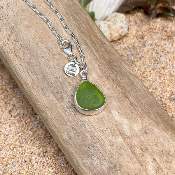 A silver chain necklace with a teardrop-shaped green sea glass pendant, resting on a piece of weathered wood over a sandy beach background.
