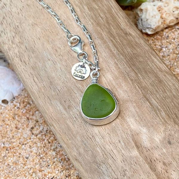 A sterling silver necklace with a green sea glass teardrop pendant, resting on a piece of driftwood over a sandy beach background.