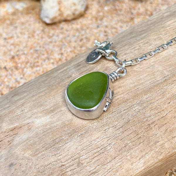 A sea glass pendant necklace with a green teardrop-shaped stone in a silver bezel setting, resting on a piece of weathered wood with sand in the background.
