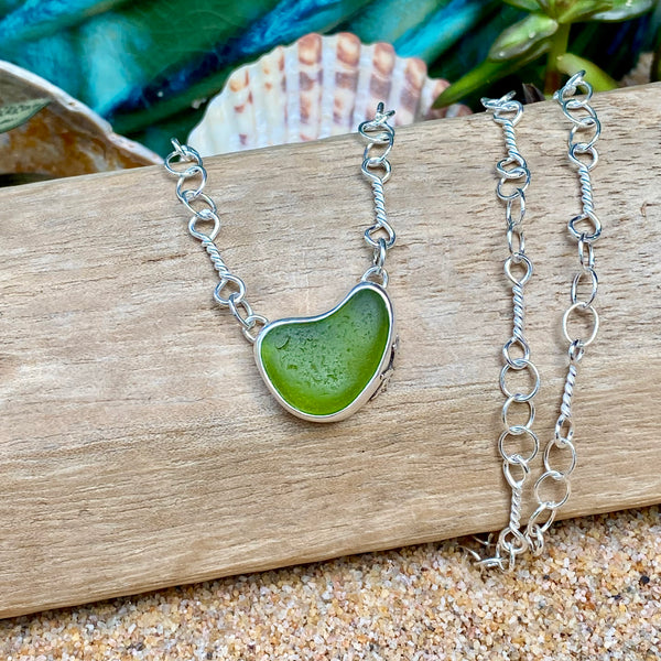 A silver chain necklace with a heart-shaped green sea glass pendant, displayed on a piece of driftwood with seashells and sand in the background.
