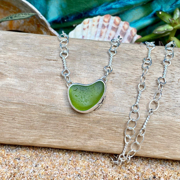 A silver chain necklace with a heart-shaped green sea glass pendant, displayed on a piece of wood with seashells and sand in the background.