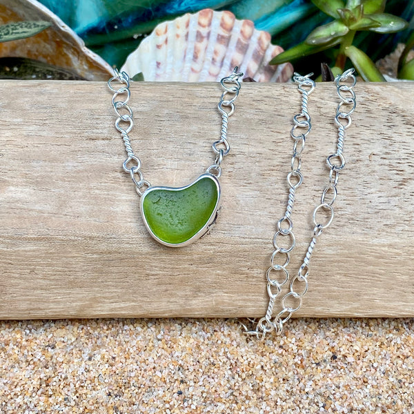 A silver chain necklace with a heart-shaped green sea glass pendant, displayed on a piece of weathered wood with seashells and sand in the background.
