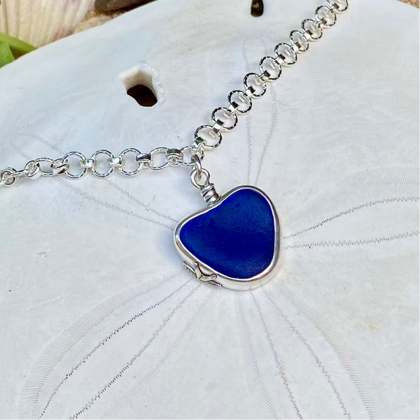 A silver heart-shaped pendant necklace with a deep blue sea glass center, displayed on a white sand dollar.