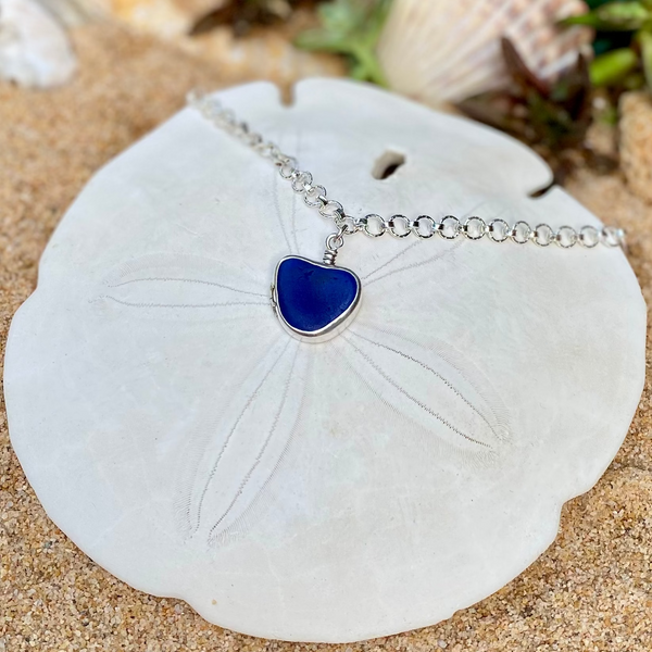 A silver chain necklace with a heart-shaped dark blue sea glass pendant, displayed on a white sand dollar shell over sand with seashells and greenery in the background.
