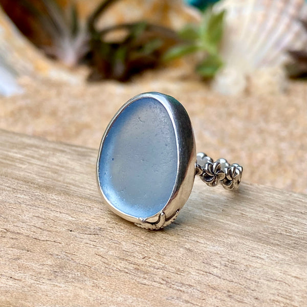 A silver ring featuring a large, smooth, oval-shaped light blue stone, displayed on a piece of weathered wood with a blurred background of shells and sand.