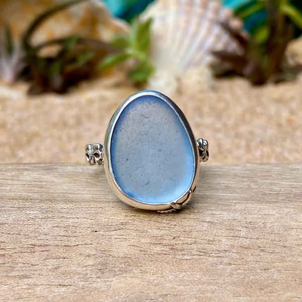 A sea glass ring featuring a large, smooth, oval-shaped light blue stone set in a silver-toned band, resting on a wooden surface with seashells and greenery blurred in the background.