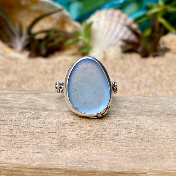 A silver ring with a large, teardrop-shaped light blue glass cabochon, displayed on a piece of wood with a sandy, shell-themed background.