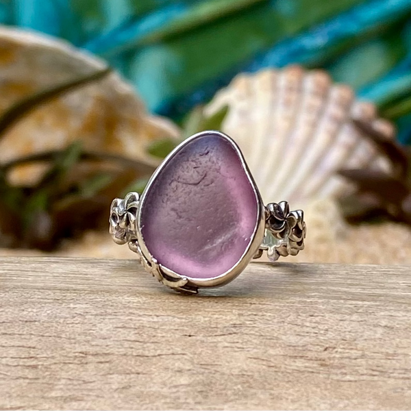 A sea glass ring featuring a smooth, teardrop-shaped lavender purple stone set in a textured silver band, displayed on a wooden surface with seashells and tropical foliage in the background.
