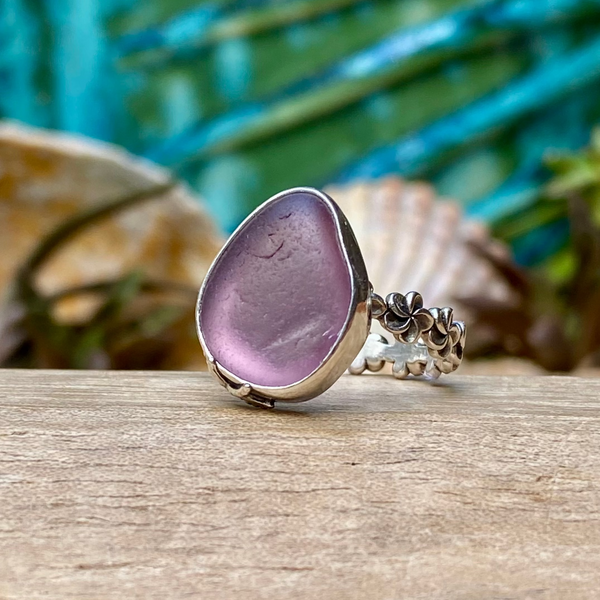 A sea glass ring with a large lavender-purple teardrop-shaped stone in a silver bezel setting, displayed on a wooden surface with seashells and a blue tropical background.