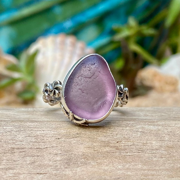 A sea glass ring featuring a teardrop-shaped lavender purple stone in a silver bezel setting with decorative floral accents on the band, resting on a wooden surface with a blurred beach-themed background.