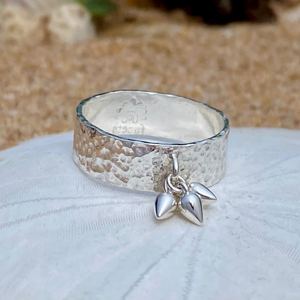 A hammered sterling silver ring with a dangling pear-shaped drops on white sand dollar with a sandy beach blurred in the background.