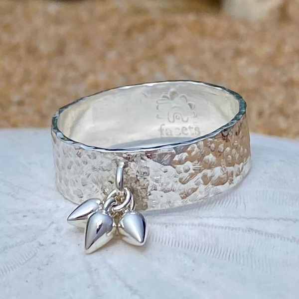 A hammered sterling silver ring with a dangling pear-shaped drops resting on a white sand dollar with a sandy beach blurred in the background.