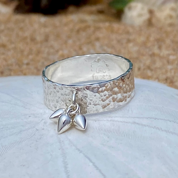 A hammered sterling silver ring with a dangling pear-shaped drops resting on a white sand dollar with a sandy beach blurred in the background.