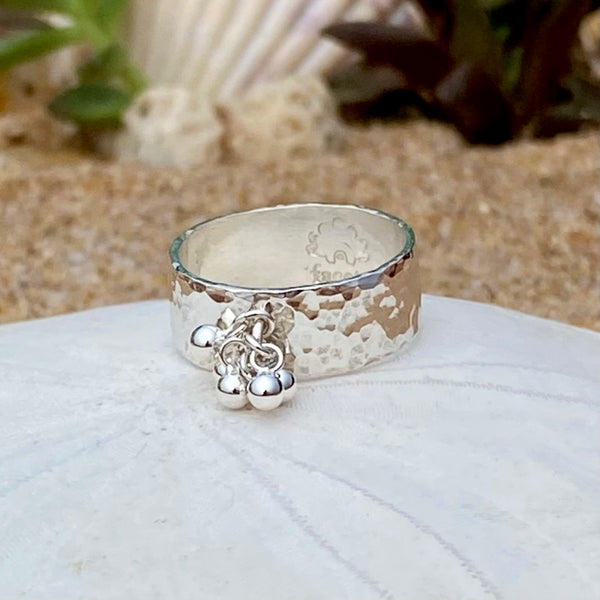 A hammered sterling silver ring with a small dangling silver balls, resting on a white sea urchin shell with a sandy beach and green plants blurred in the background.