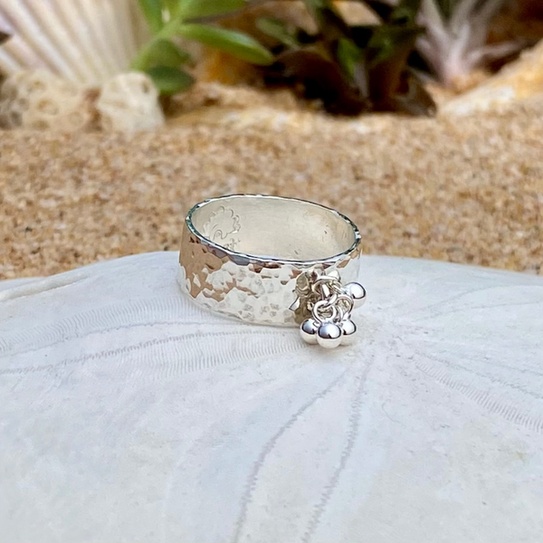 A hammered sterling silver ring with a small dangling silver balls, resting on a white sea urchin shell with a sandy beach and green plants blurred in the background.
