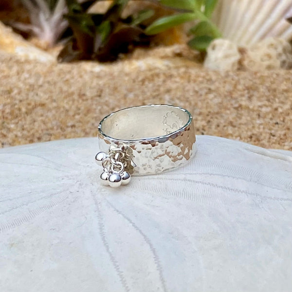 A hammered sterling silver ring with a small dangling silver balls, resting on a white sea urchin shell with a sandy beach and green plants blurred in the background.