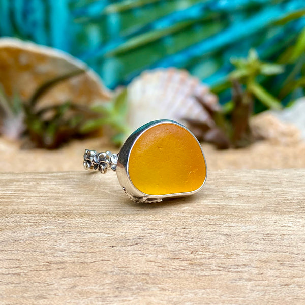 A sea glass ring featuring a smooth, polished orange-yellow stone in a silver bezel setting, displayed on a wooden surface with seashells and tropical foliage in the background.