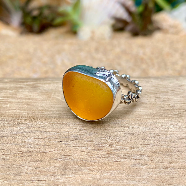 A handmade ring featuring a large, polished orange-yellow stone with a silver bezel and a silver beaded band, resting on a wooden surface with a blurred natural background.