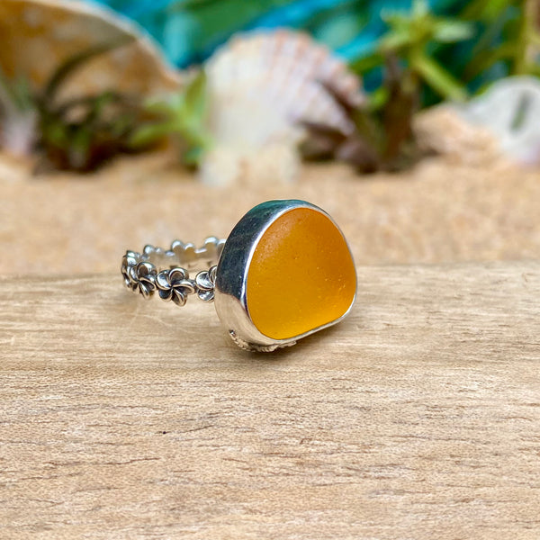 A sea glass ring featuring a large, irregularly shaped golden-orange stone set in a silver band with small floral details, resting on a wooden surface with a blurred beach background.