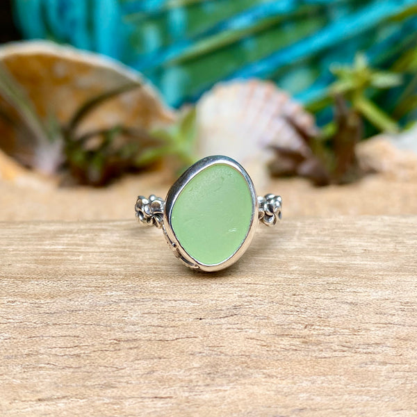 A sea glass ring featuring a smooth, rounded light green stone in a silver bezel setting, displayed on a wooden surface with blurred seashells and tropical foliage in the background.