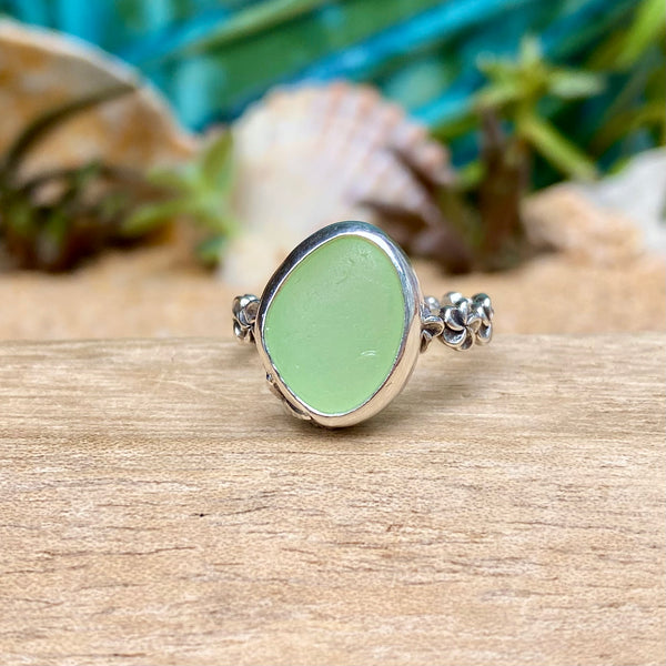 A silver ring with an irregular, rounded light green stone, resting on a wooden surface with seashells and green foliage blurred in the background.