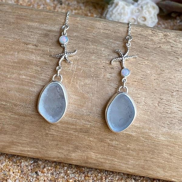 A pair of silver threader earrings with sea star detail, small iridescent opal cabochons and teardrop-shaped ice blue sea glass, displayed on a piece of weathered wood over sand.