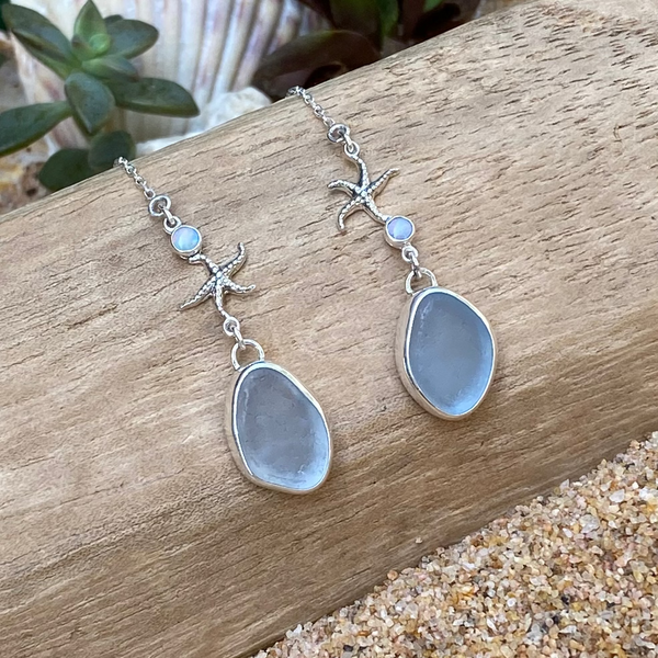 A pair of silver threader earrings with sea star detail, small iridescent opal cabochons and teardrop-shaped ice blue sea glass, displayed on a piece of weathered wood over sand.