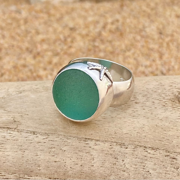 A sterling silver signet ring with a round, frosty teal sea glass marble and a small starfish detail on the band, resting on a wooden surface with a sandy background.