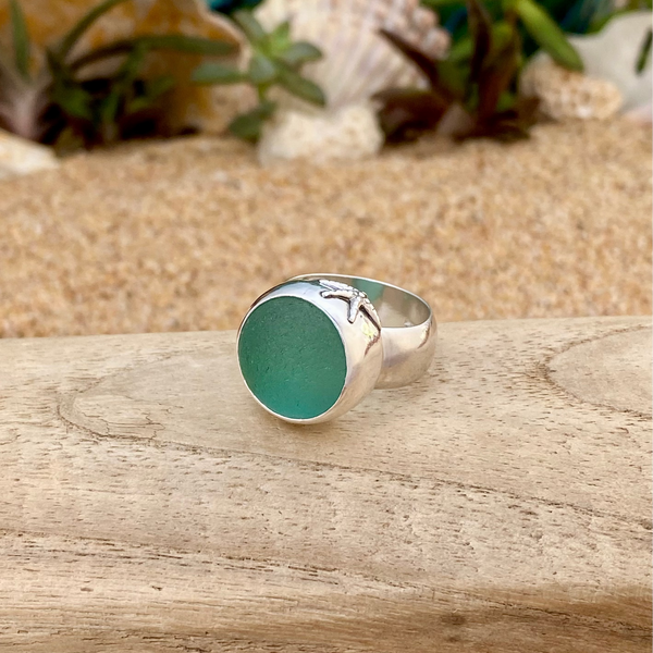 A sterling silver signet ring with a round, frosty teal sea glass marble and a small starfish detail on the band, resting on a wooden surface with a sandy background.