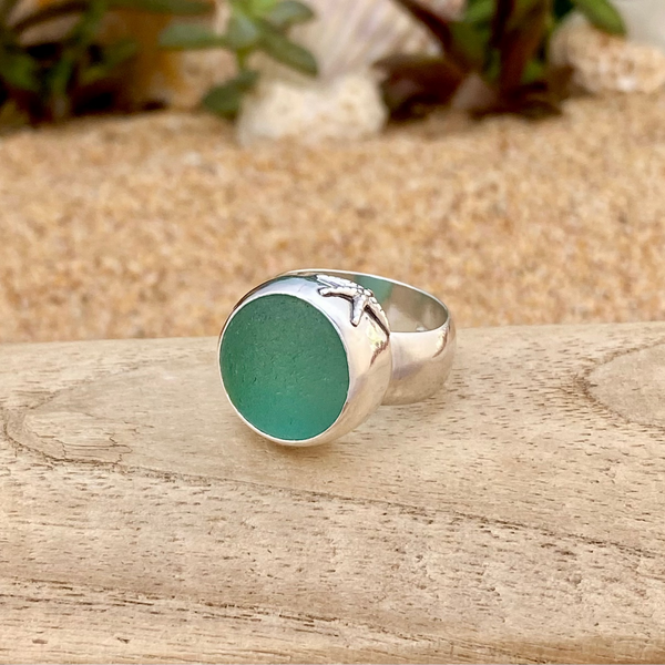 A sterling silver signet ring with a round, frosty teal sea glass marble and a small starfish detail on the band, resting on a wooden surface with a sandy background.
