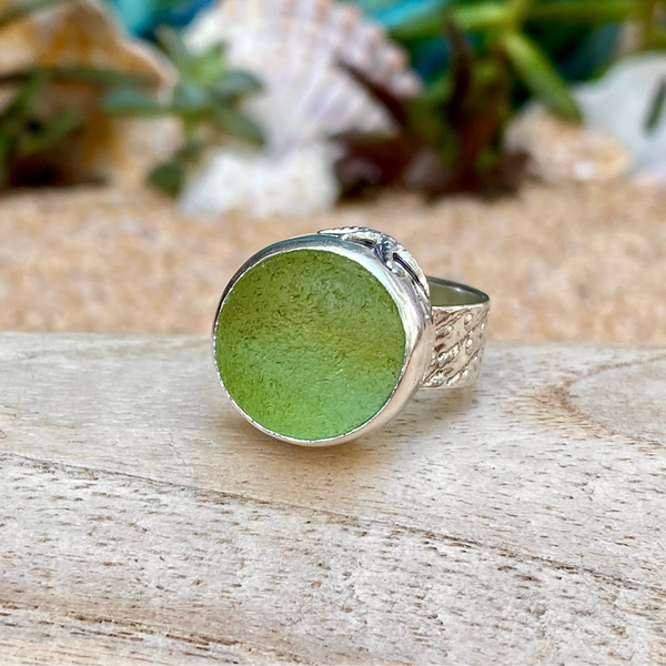 A sterling silver ring with a large, round, frosty green sea glass marble, resting on a light-colored wooden surface with a sandy background.