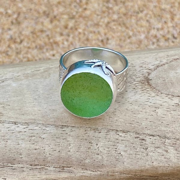 A sterling silver ring with a large, round, frosty green sea glass marble, resting on a light-colored wooden surface with a sandy background.