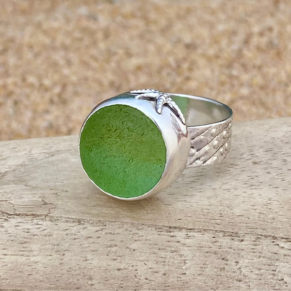 A sterling silver ring with a large, round, frosty green sea glass marble, resting on a light-colored wooden surface with a sandy background.