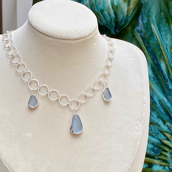 A sterling silver necklace with circular links and three teardrop-shaped sea glass pendants, displayed on a cream-colored jewelry bust. The background features a large green seashell and small seashells on a wooden surface.