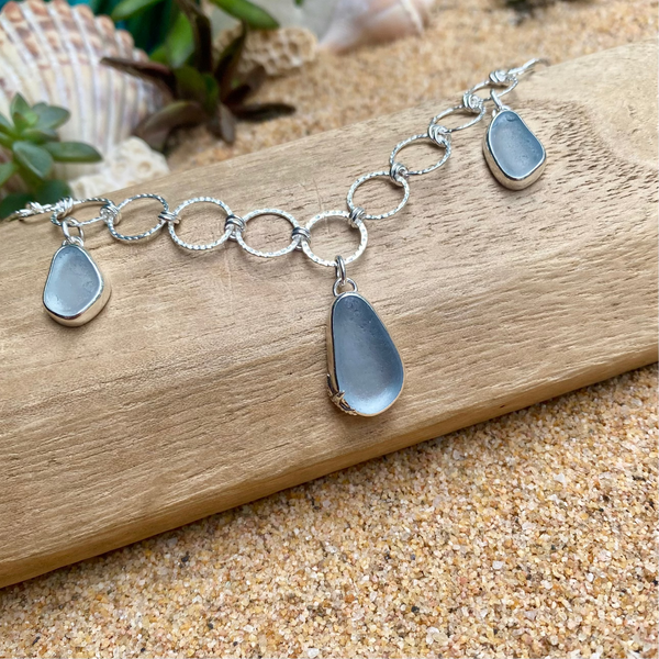 A silver circular link necklace with three teardrop-shaped sea glass pendants in a muted ice blue color, displayed on a piece of wood over sand with seashells in the background.