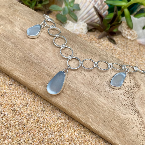 A silver chain bracelet with three teardrop-shaped sea glass pendants in a muted blue-gray color, displayed on a piece of wood over sand with seashells in the background.