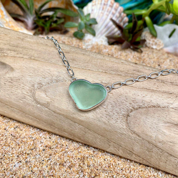 A heart-shaped sea glass pendant necklace in a silver bezel setting, resting on a piece of driftwood over sand with seashells and greenery in the background.