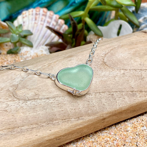 A heart-shaped sea glass pendant necklace in a silver bezel setting, resting on a piece of driftwood over sand with seashells and greenery in the background.