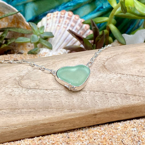 A heart-shaped sea glass pendant necklace in a silver bezel setting, resting on a piece of driftwood over sand with seashells and greenery in the background.