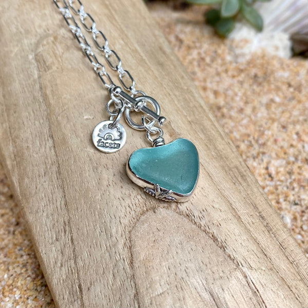 A sterling silver necklace with a heart-shaped sea glass pendant in a muted teal-blue color, resting on a piece of weathered wood over sand.