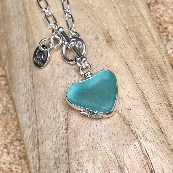 A sterling silver chain front toggle clasp necklace with a heart-shaped turquoise colored sea glass pendant and a small dangling tag, displayed on a piece of weathered wood over sand.