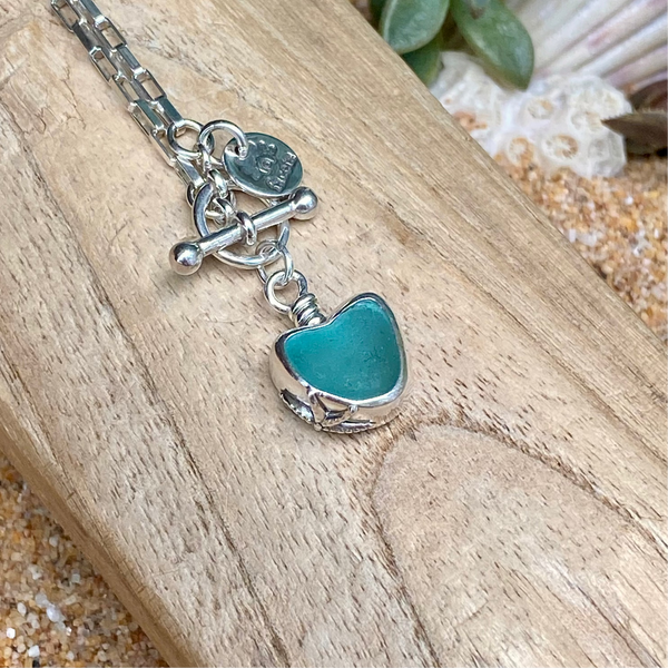 A sterling silver chain necklace with a toggle clasp and a heart-shaped teal green sea glass pendant, displayed on a piece of wood with sand and seashells in the background.
