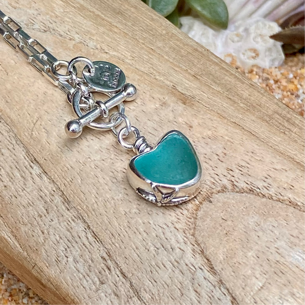 A sterling silver necklace with a heart-shaped teal green sea glass pendant, resting on a light wood surface with seashells and a succulent in the background.