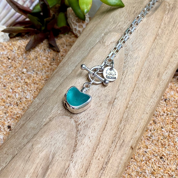 A sterling silver chain necklace with a toggle clasp and a heart-shaped teal green sea glass pendant, displayed on a piece of wood over sand with small green leaves in the background.